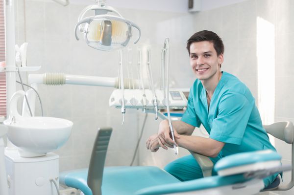 دندان های سالم و لبخند دکتر جدی مستقیم به دوربین دندانپزشکی که روی صندلی دندانپزشکی نشسته و مته دندانپزشکی را در دست گرفته است دندانپزشک دست هایش را به صورت ضربدری روی هم گذاشت
