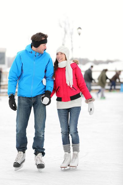 زوج جوان رمانتیک با لباس گرم زمستانی که دست در دست هم گرفته و در حال اسکیت روی یخ به یکدیگر لبخند می زنند