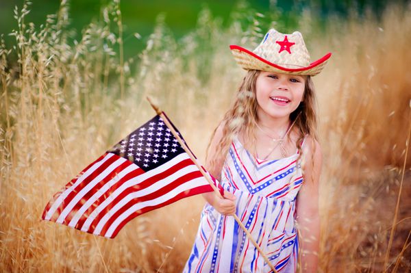 دختر کوچولوی شایان ستایش لبخند می زند و پرچم آمریکا را بیرون تکان می دهد لباسش با نوار و ستاره کلاه کابوی کودک خندان چهارم ژوئیه - روز استقلال را جشن می گیرد