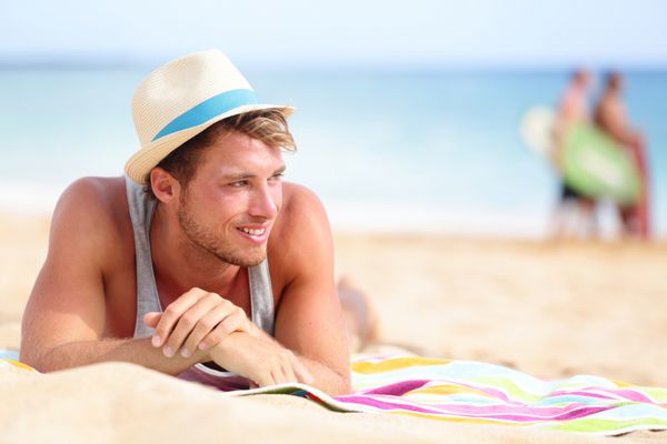مردی در ساحل دراز کشیده در شن و به طرفین نگاه می کند و خوشحال است که کلاه تابستانی هیپستر بر سر دارد مدل مرد جوان در حال لذت بردن از تعطیلات سفر تابستانی در کنار اقیانوس