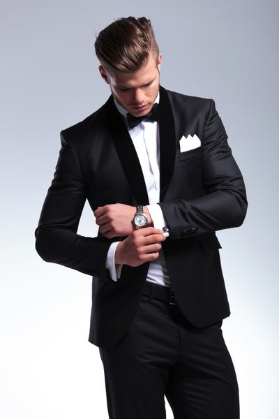 مرد جوان شیک پوش با لباس تاکسیدو در حالی که دکمه سرآستین خود را درست می کند به آنها نگاه می کند در زمینه خاکستری