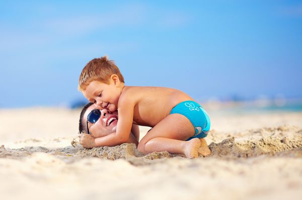 بچه شادی که به آرامی سر پدرش را در شن های ساحل بغل کرده است