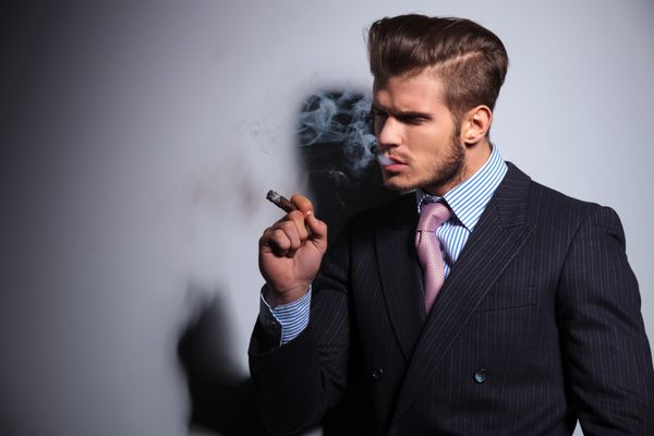 نمای کناری یک مدل مد با کت و شلوار و کراوات در حال لذت بردن از سیگار خود در زمینه خاکستری