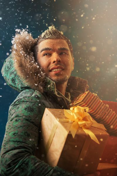 تصویر روشن مرد خوش تیپ در حال حمل هدایای کریسمس در فضای باز در زمستان در شب نور طلایی و زرق و برق جادویی در اطراف او