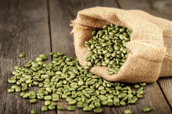 دانه های قهوه سبز در کیسه قهوه ساخته شده از کرفس روی سطح چوبی در وسط قاب فوکوس شده است