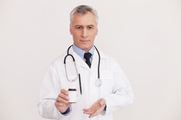 ارائه داروهای جدید دکتر ارشد موهای خاکستری با لباس فرم به شیشه دارو اشاره می کند و در حالی که ایزوله روی سفید ایستاده به دوربین نگاه می کند