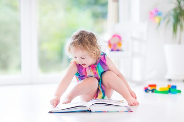 دختر نوپای مجعد ناز در حال خواندن کتاب در اتاق خواب آفتابگیر با پنجره های بزرگ با دید باغ با اسباب بازی های اطرافش نشسته روی زمین