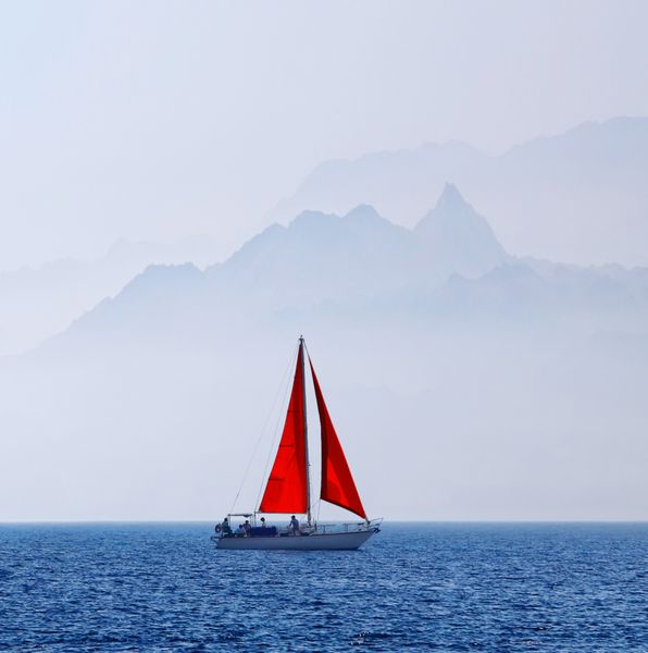 قایق تفریحی با بادبان قرمز در پس زمینه کوه در دریا