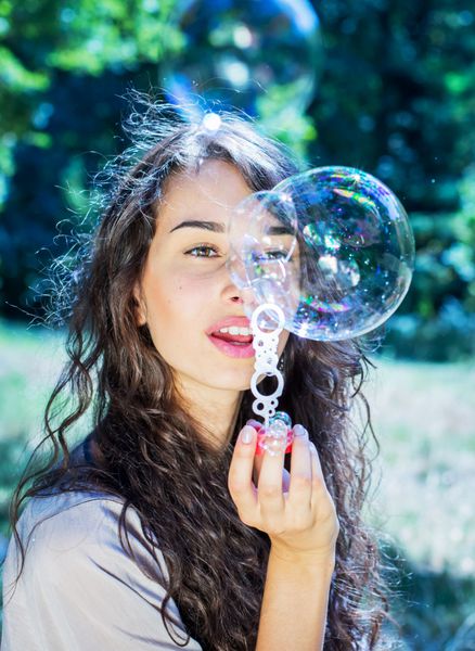 دختر جوان رمانتیک در حال باد کردن حباب های صابون رنگارنگ در پارک