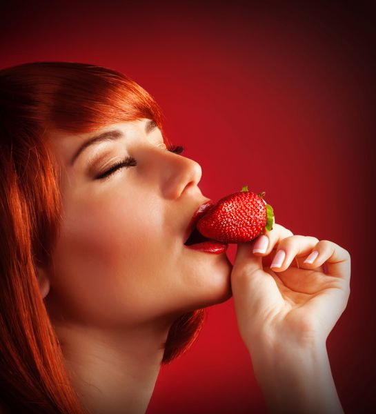 عکس زن اغوا کننده در حال خوردن توت فرنگی پرتره نزدیک از زن شهوانی مو قرمز در حال گاز گرفتن میوه های آبدار جدا شده در پس زمینه قرمز رژیم میوه ای دسر خوشمزه روز ولنتاین مفهوم لذت