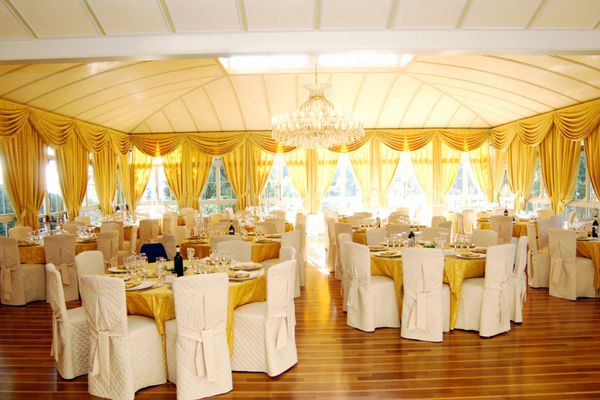 فضای داخلی مجلل عروسی یا محل برگزاری مراسم با میزها و چیدمان میزهای برجسته با طلایی غنی و پنجره‌های متعدد که نوری روشن و مطبوع می‌دهد