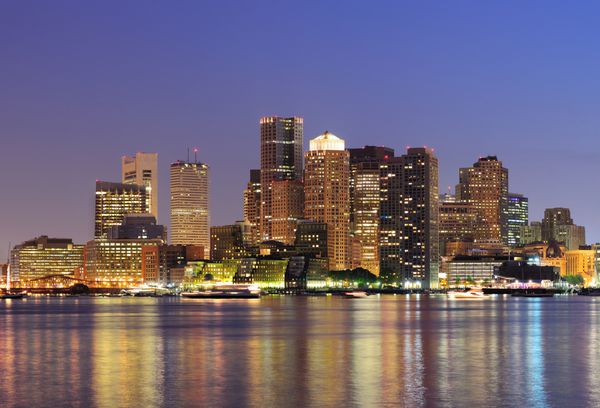 منظره‌ای از خط افق مرکز شهر بوستون با آسمان‌خراش‌هایی بر فراز آب با انعکاس‌هایی در غروب که با نورها روشن شده‌اند