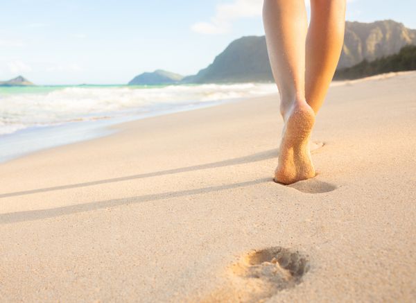 سفر در ساحل - زنی که در ساحل شنی قدم می زند و رد پایی را در شن ها می گذارد جزئیات نزدیک از پاهای زن و ماسه طلایی در ساحل در هاوایی