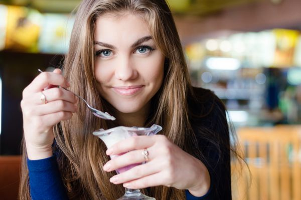 زن جوان زیبا و شاد با لبخند به دوربین نگاه می کند که در رستوران یا کافه نشسته است و پرتره نزدیک بستنی می خورد لبخند می زند و در دوربین نگاه می کند