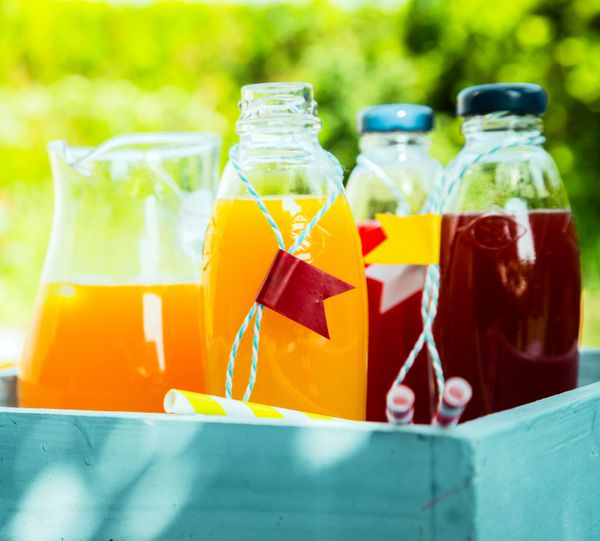 آب پرتقال و توت تازه سالم خانگی در بطری های شیشه ای و کوزه ها در یک جعبه فیروزه ای چوبی روی میز در باغ تابستانی ایستاده است تا یک نوشیدنی طراوت بخش باشد