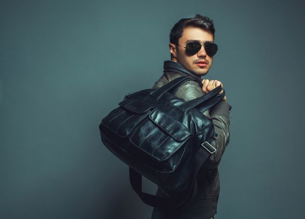 پرتره مرد جوان خوش تیپ مد با کیف چرمی که عینک آفتابی و ژاکت چرمی بر تن دارد نزدیک فضای کپی شات استودیویی