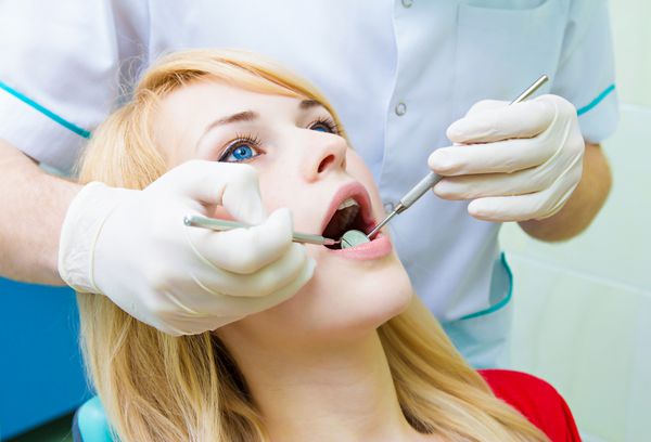 نمای نزدیک مجسمه زن جوان زن بلوند زیبا بیمار نشسته روی صندلی دندانپزشکی مطب با دهان باز و معاینه کامل دهان و دندان توسط پزشک با دستکش با استفاده از آینه