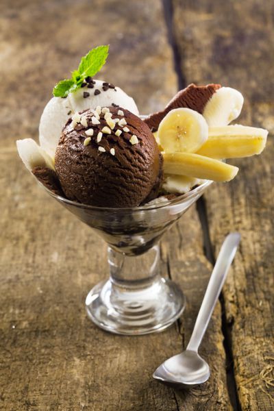 دسر خوشمزه موز و بستنی با میوه های برش خورده با قاشق های شکلات و بستنی وانیلی روی میز چوبی روستایی سرو می شود