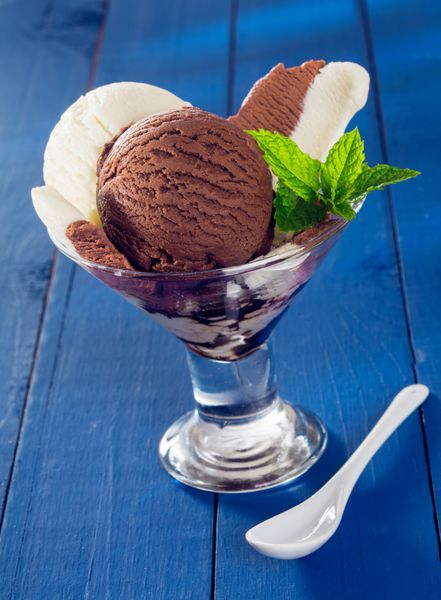 بستنی شکلاتی و وانیلی در یک ظرف شیشه ای مخروطی شکل روی تخته های چوبی آبی رنگ شده سرو می شود