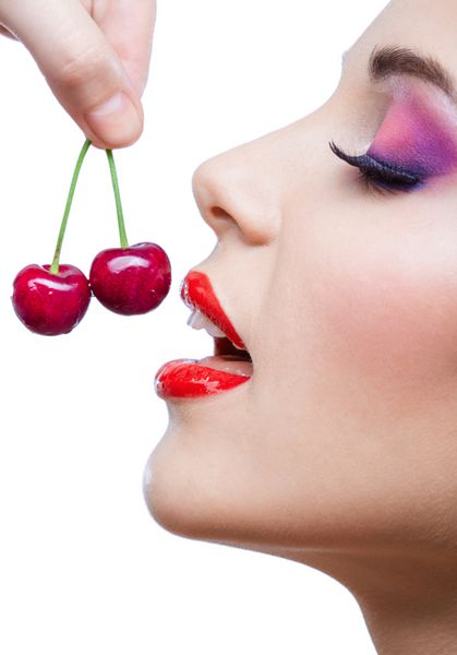 از نزدیک دختری با لب‌های قرمز و آرایش روشن در حال خوردن دو توت جدا شده روی سفید مفهوم شهوانی و سلامتی