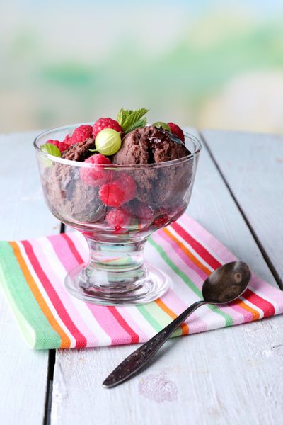 بستنی شکلاتی با برگ نعنا و توت های رسیده در کاسه شیشه ای روی میز چوبی رنگی در پس زمینه روشن