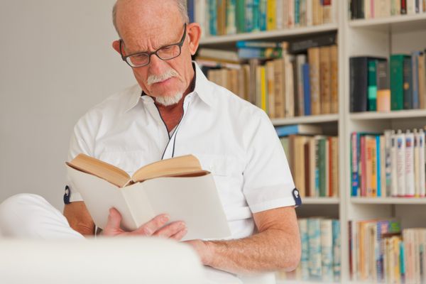مرد ارشد با عینک در حال خواندن کتاب جلوی قفسه کتاب