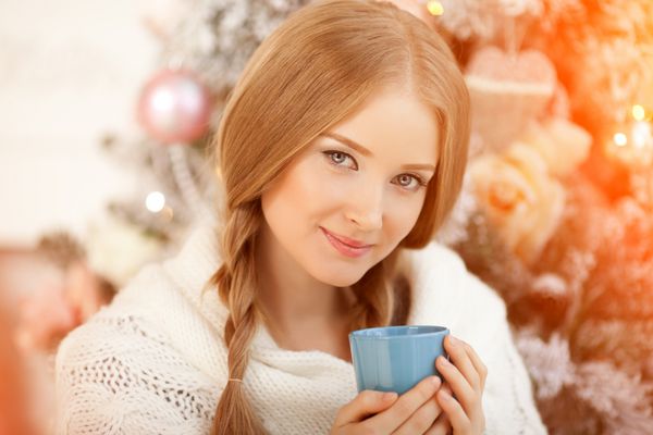 زن جوان زیبا در حال نوشیدن چای در درخت کریسمس دختر زیبا کریسمس را با یک فنجان کاکائو جشن می گیرد
