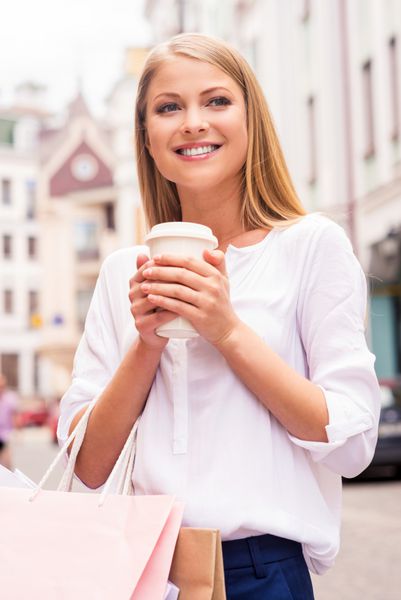 تازه شدن قبل از فروشگاه بعدی زن جوان و خندان زیبا که کیسه‌های خرید و فنجان نوشیدنی داغ در دست دارد در حالی که بیرون از خانه ایستاده است