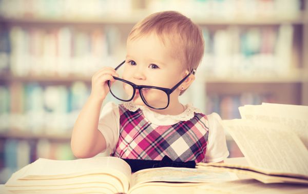 دختر بچه خنده دار شاد با عینک در حال خواندن کتاب در کتابخانه