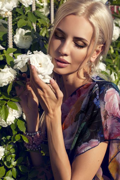 پرتره زنی زیبا که یک گل رز سفید بزرگ را در باغ تابستانی بو می کند