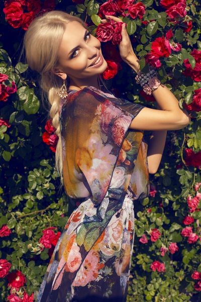 زن زیبا لبخند با موهای بلوند در کنار دیواری از گل رز عکس می گیرد