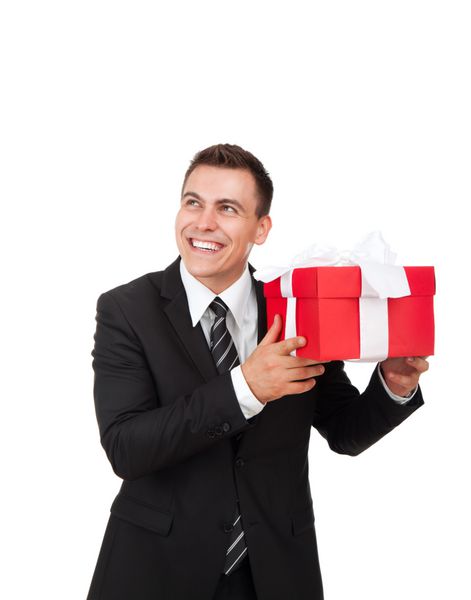 تاجر هیجان زده لبخند شاد جعبه هدیه را در دست بگیرید مرد تاجر جعبه هدیه قرمز رنگ با کمان روبانی جدا شده روی پس زمینه سفید ارائه می دهد