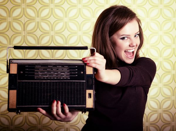 پرتره هنری زن جوان خندان و شادی که پخش کننده رادیو را در اتاق با کاغذ دیواری قدیمی سبک‌سازی رترو دهه 60 تا 70 در دست دارد
