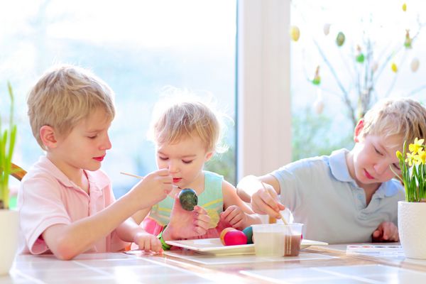 گروهی از کودکان ناز از یک خانواده دو برادر دوقلو و خواهر کوچکترشان در حال تزئین و نقاشی تخم مرغ های عید پاک در یک روز آفتابی در آشپزخانه نشسته اند تمرکز انتخابی روی دختر
