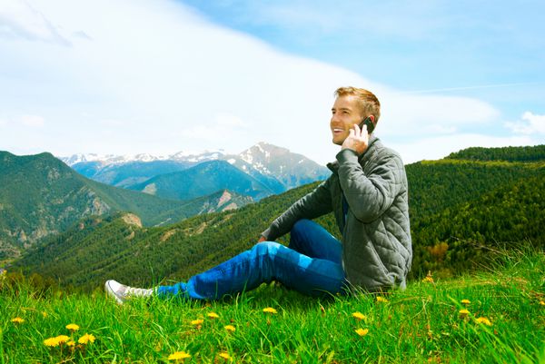 مرد جوان خوش تیپ در حال تماس با تلفن همراه خود که در چمن در زمین سبز نشسته است مرد گاه به گاه در حال صحبت با تلفن همراه در فضای باز کوه ها مفهوم ارتباط آزادی تعطیلات فن آوری