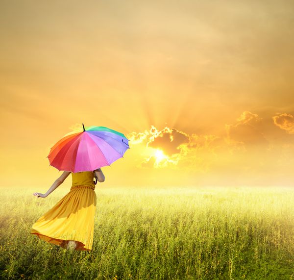 زن زیبا که چتر رنگارنگ را در زمین چمن سبز و غروب خورشید در دست دارد
