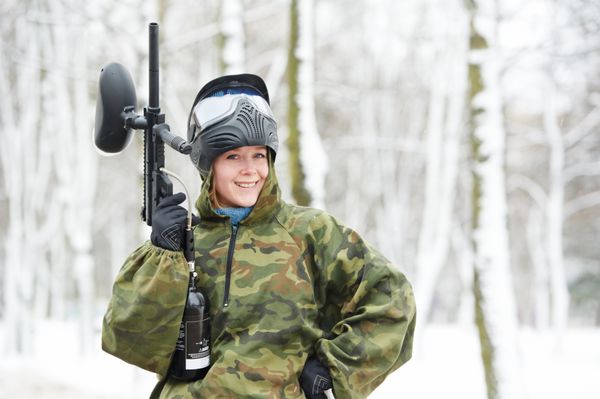 بازیکن شاد زن پینت بال ورزش افراطی با پوشیدن ماسک محافظ و لباس استتار با تفنگ نشانگر در زمستان در فضای باز