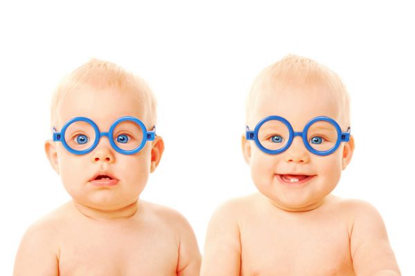 دو نوزاد پسر دوقلو با عینک یکی از بچه ها جدی است بچه دیگر لبخند می زند دانش آموزان جوان جدا شده در زمینه سفید