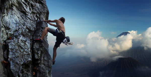 مرد جوانی در حال بالا رفتن از دیوار صخره ای طبیعی با آتشفشان ها در پس زمینه
