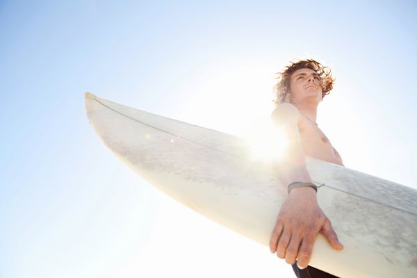 مرد ورزشکار جوان جذاب موج سواری که لباس غواصی لاستیکی نئوپرن به تن دارد و در حالی که تخته موج سواری خود را در یک روز آفتابی در برابر آسمان آبی شدید حمل می کند به افق نگاه می کند