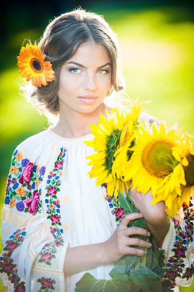 دختر جوانی که بلوز سنتی رومانیایی پوشیده است و عکس بیرون از گل آفتابگردان را در دست دارد پرتره دختر بلوند زیبا با دسته گلهای زرد روشن زن زیبا با گل زرد در حالت ژست مو