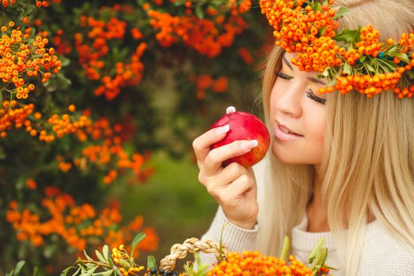 پرتره زن خندان با پوشیدن لوازم جانبی پشمی زن تاج گل نارنجی با سیب قرمز در دست پرتره زن جوان زیبا که در پاییز در فضای باز قدم می زند