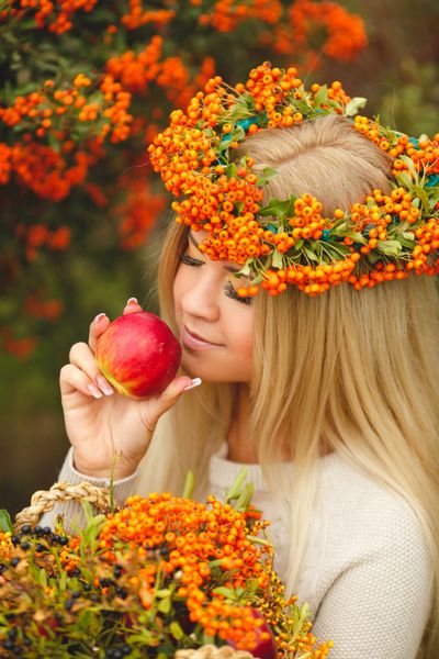 پرتره زن خندان با پوشیدن لوازم جانبی پشمی زن تاج گل نارنجی با سیب قرمز در دست پرتره زن جوان زیبا که در پاییز در فضای باز قدم می زند