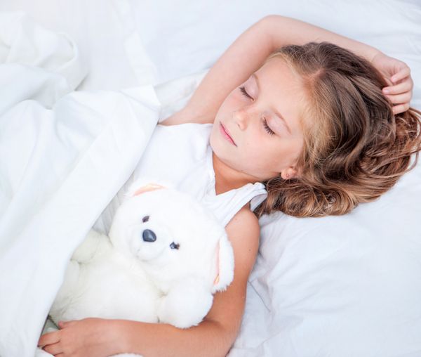 دختر زیبای شش ساله ای که با خرس عروسکی خود روی تخت خوابیده است