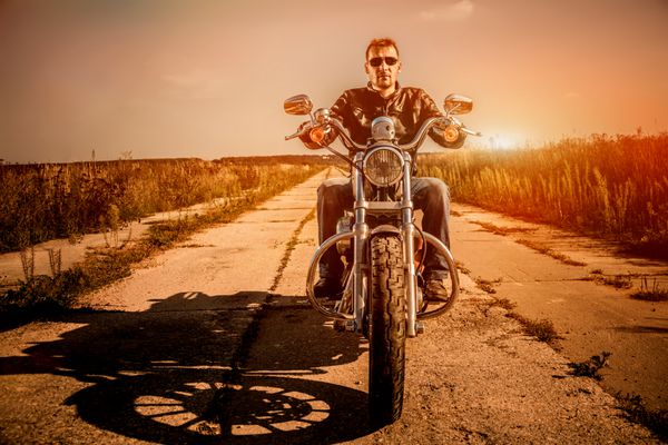 مرد دوچرخه‌سواری با یک ژاکت چرمی و عینک آفتابی که روی موتور سیکلت خود نشسته و به غروب آفتاب نگاه می‌کند
