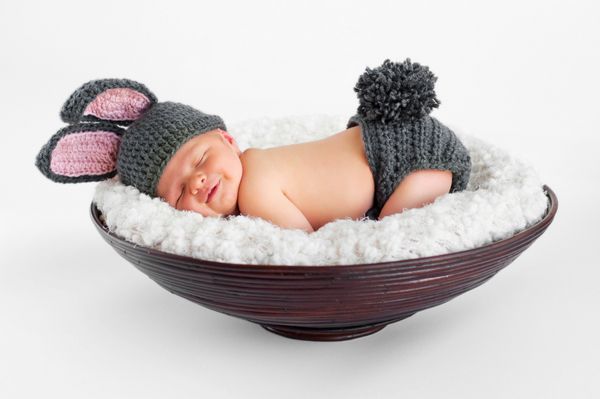 نوزاد پسر هشت روزه خندان با گوش های خرگوش و پوشک دم خرگوش او روی شکم در یک سبد خوابیده است در استودیو روی یک پس‌زمینه سفید جدا شده