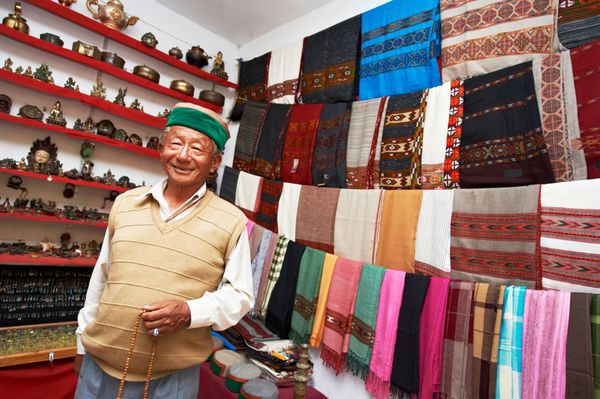 مرد هندی صاحب مغازه کوچکی که در فروشگاه خود شال لباس و سوغاتی می فروشد