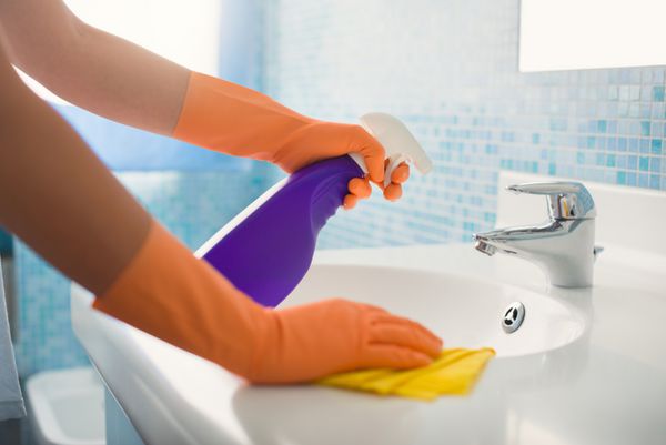 زن در حال انجام کارهای خانه در حمام تمیز کردن سینک و شیر آب با اسپری شوینده نمای برش خورده