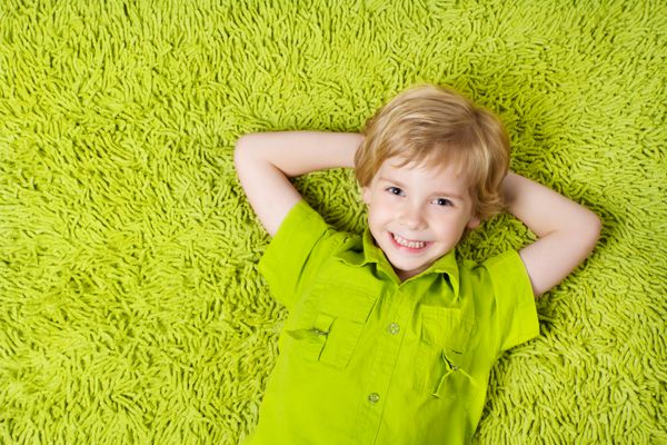 کودک شادی که روی پس زمینه فرش سبز دراز کشیده است پسری که لبخند می زند و به دوربین نگاه می کند