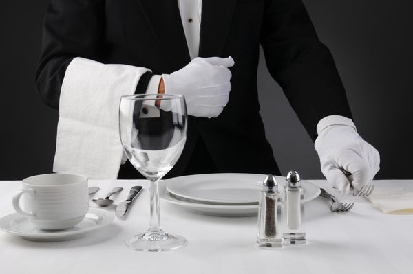 نمای نزدیک از یک پیشخدمت با لباس تاکسیدو در حال چیدن میز شام رسمی عمق میدان کم در فرمت افقی در زمینه خاکستری روشن تا تیره انسان غیرقابل تشخیص است